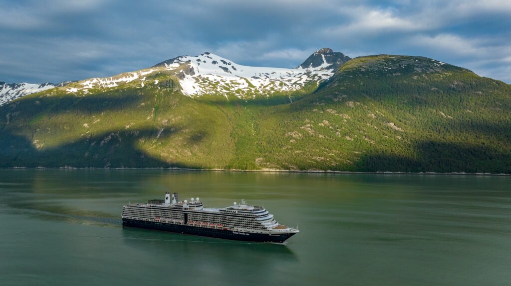Nieuw Amsterdam cruise ship sails by snow-capped mountains and green forests.